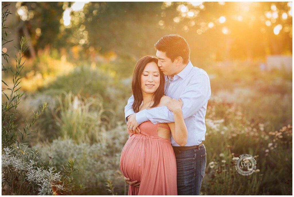 Expecting mom to be poses for maternity pictures during sunset being kissed by her husband. 